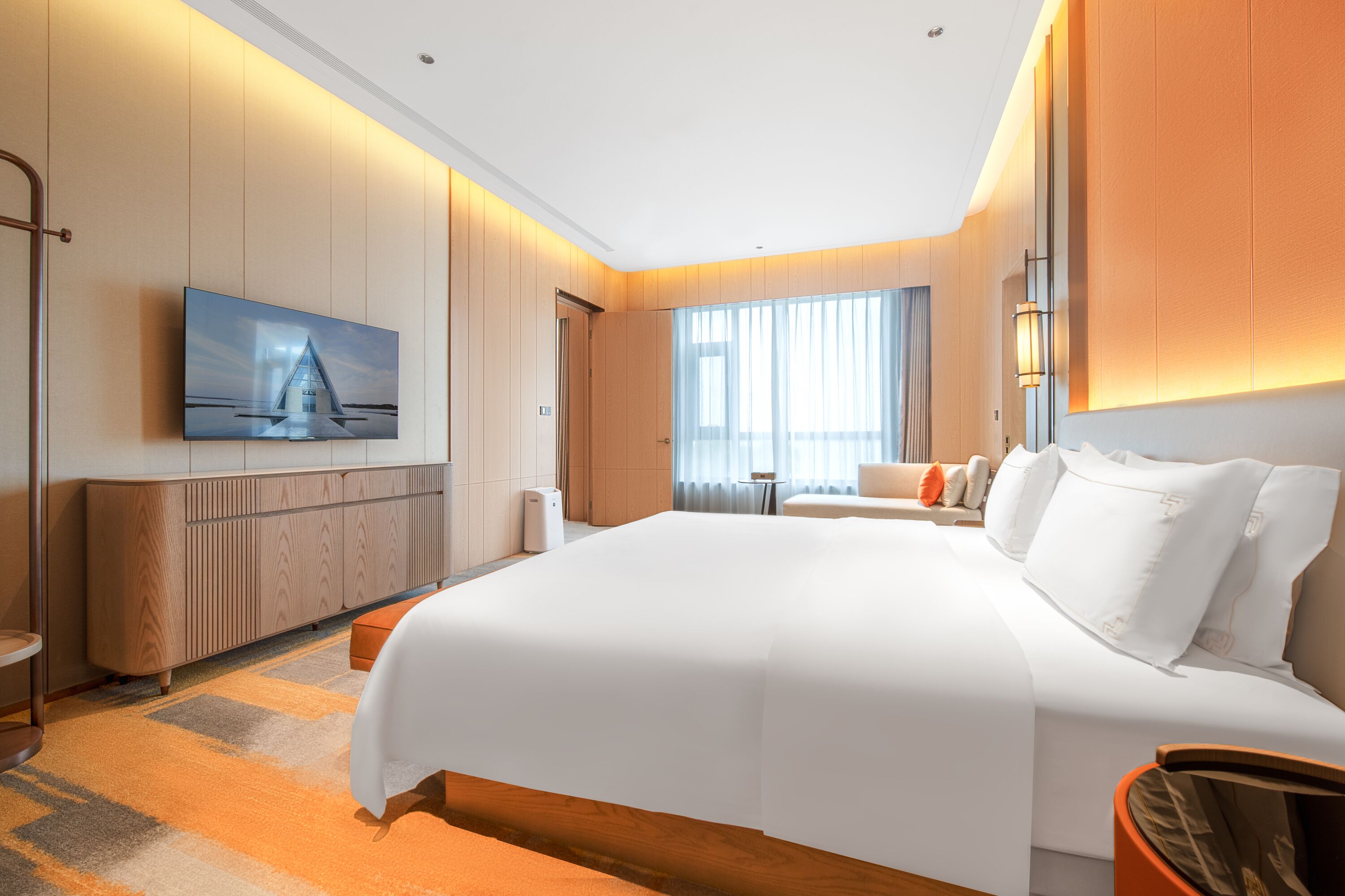 Una habitación de hotel moderna con una cama grande, un televisor de pantalla plana en la pared, un área de estar con sofá y sillones, y un ventanal con cortinas.