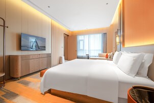 Una habitación de hotel moderna con una cama grande, un televisor de pantalla plana en la pared, un área de estar con sofá y sillones, y un ventanal con cortinas.