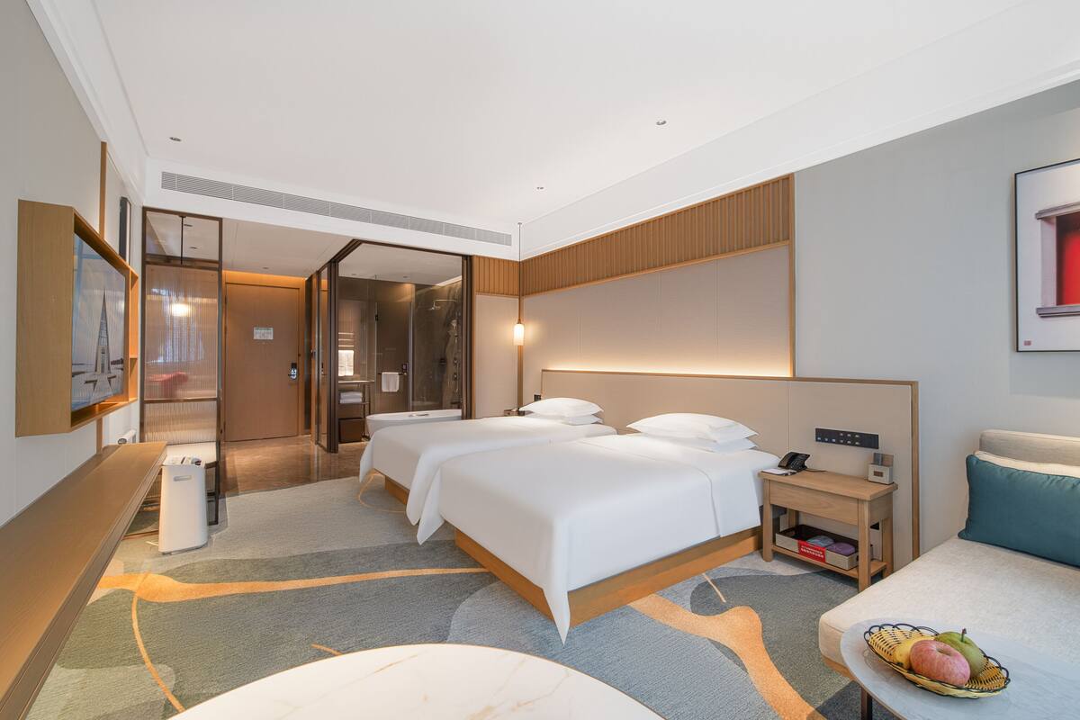 A hotel room with two beds, a desk, a chair, a small table with fruit, and a bathroom visible through a glass door.