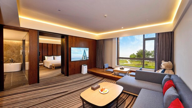 Una habitación de hotel moderna con una cama grande, una zona de estar con un sofá y una mesa, un televisor montado en la pared y vistas al exterior a través de un ventanal amplio.