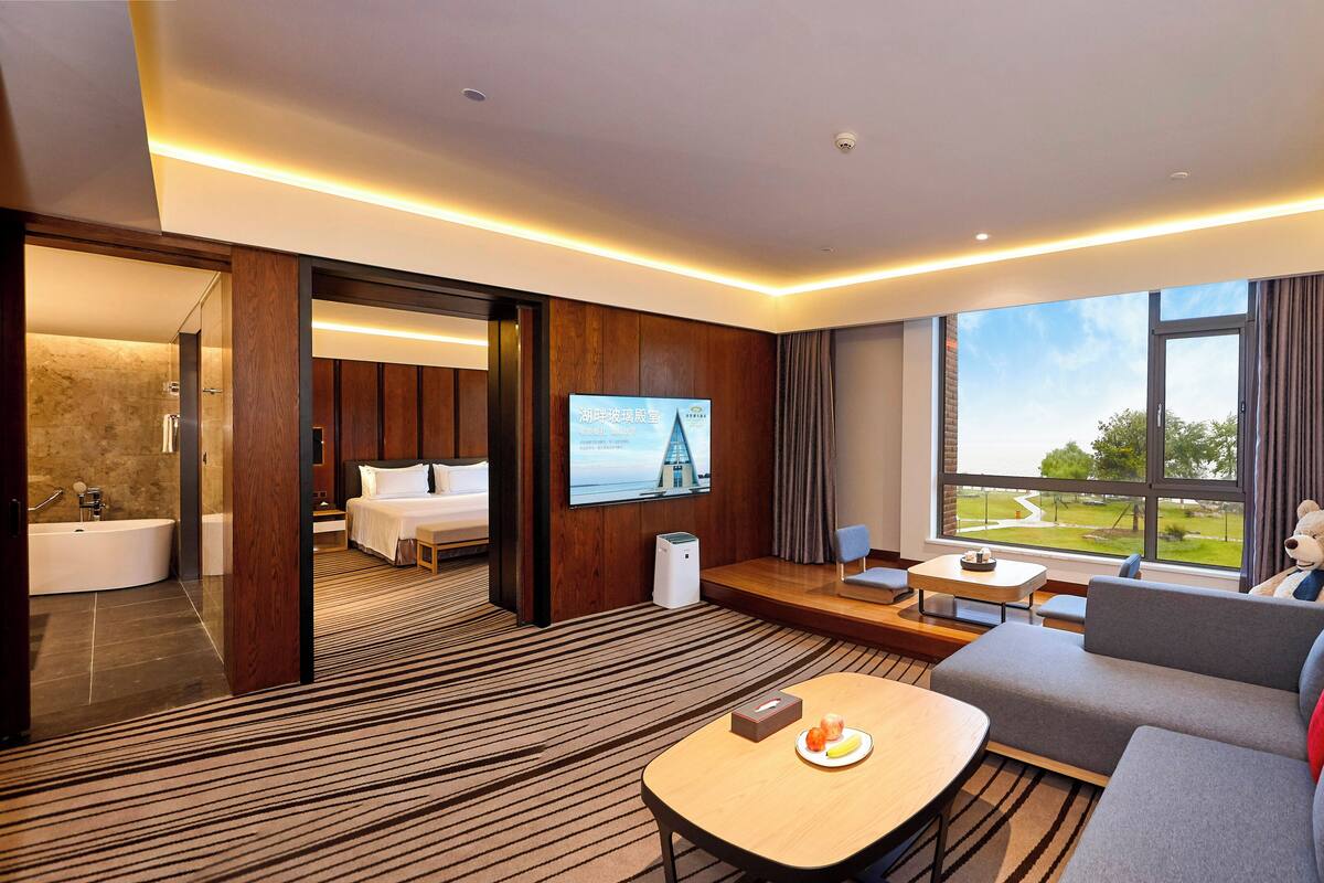 A modern hotel room with a large bed, a sitting area with a sofa and a table, a TV mounted on the wall, and a view of the outdoors through a large window.
