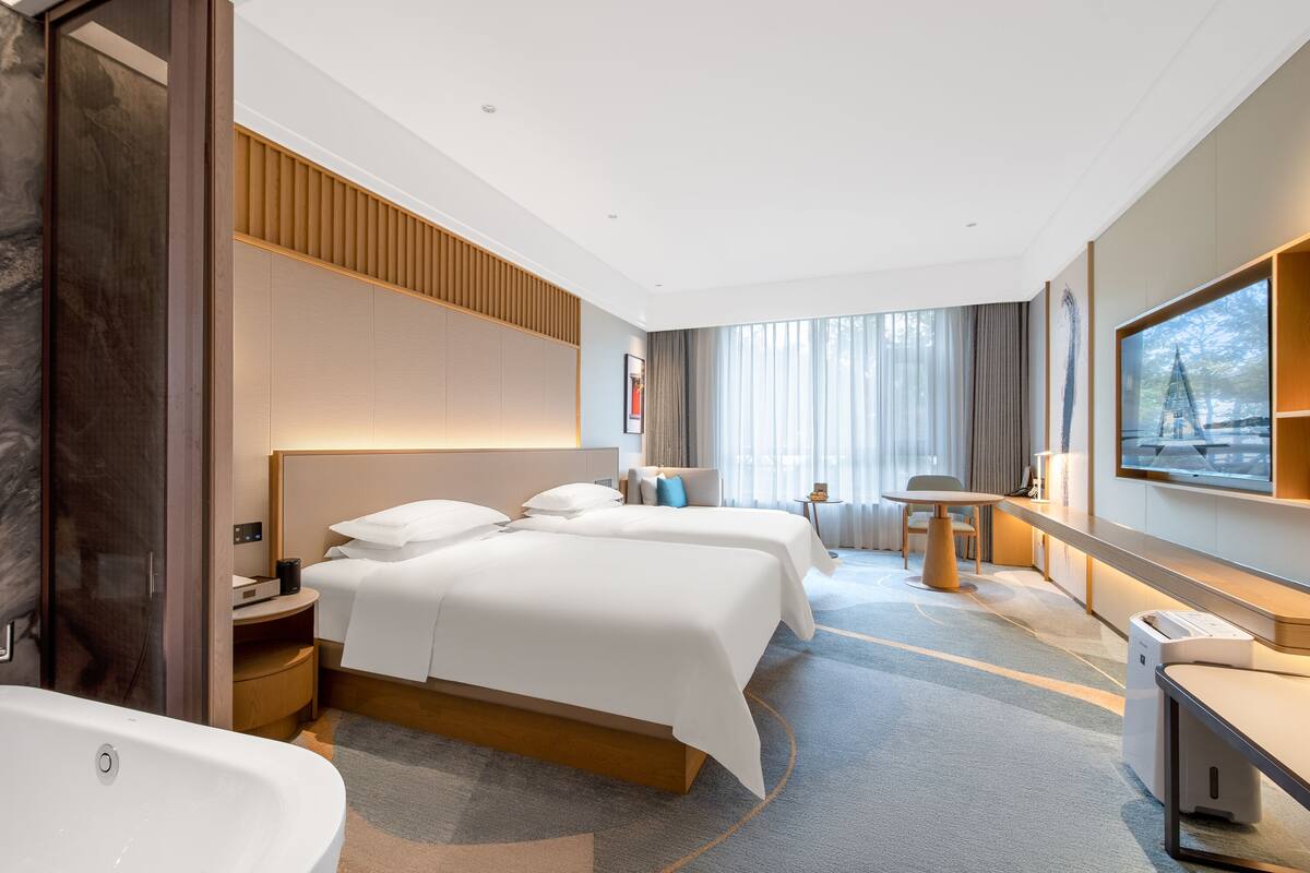 A modern hotel room with a large bed, a separate seating area, a desk, and a large window with curtains.