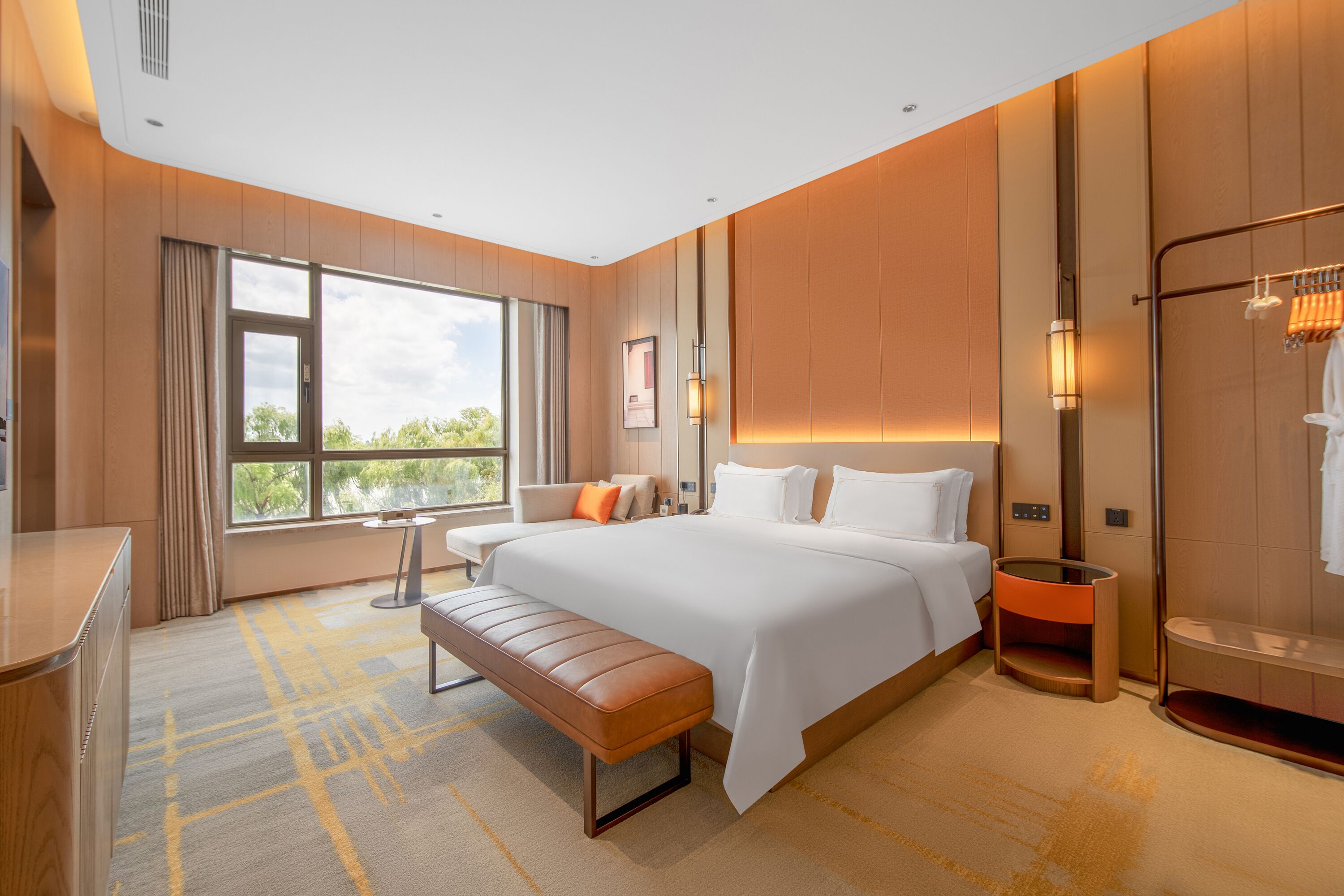 Habitación de hotel moderna con una cama grande, un banco, un escritorio y vistas al exterior.