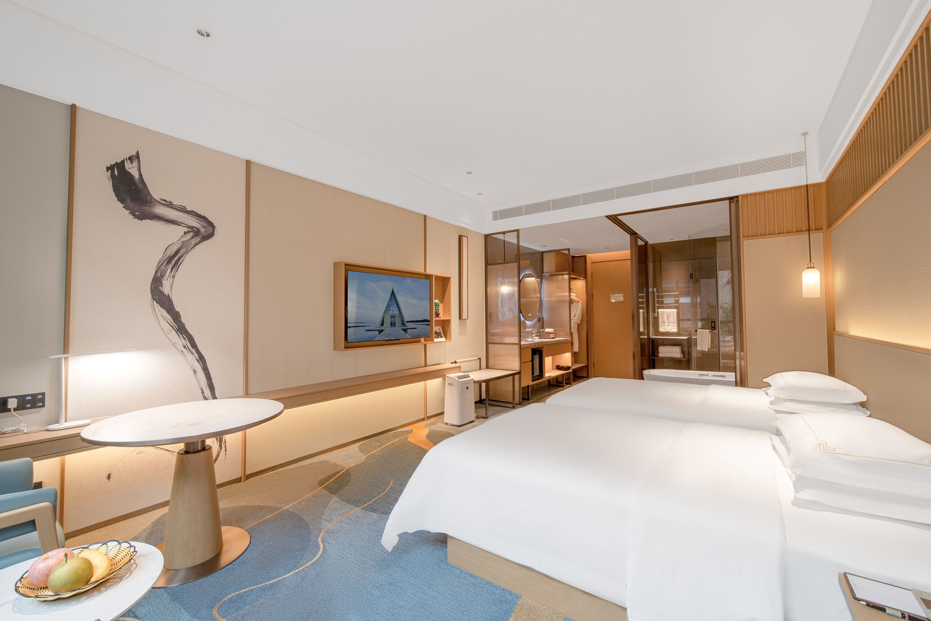 Habitación de hotel moderna con una cama grande, un escritorio, una silla, una mesita con fruta, un cuadro y un baño con puerta de vidrio.