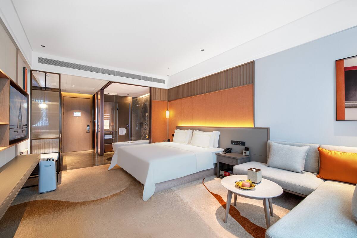 A modern hotel room with a large bed, a sofa, a small table with a fruit bowl, a desk with a lamp, and a TV.