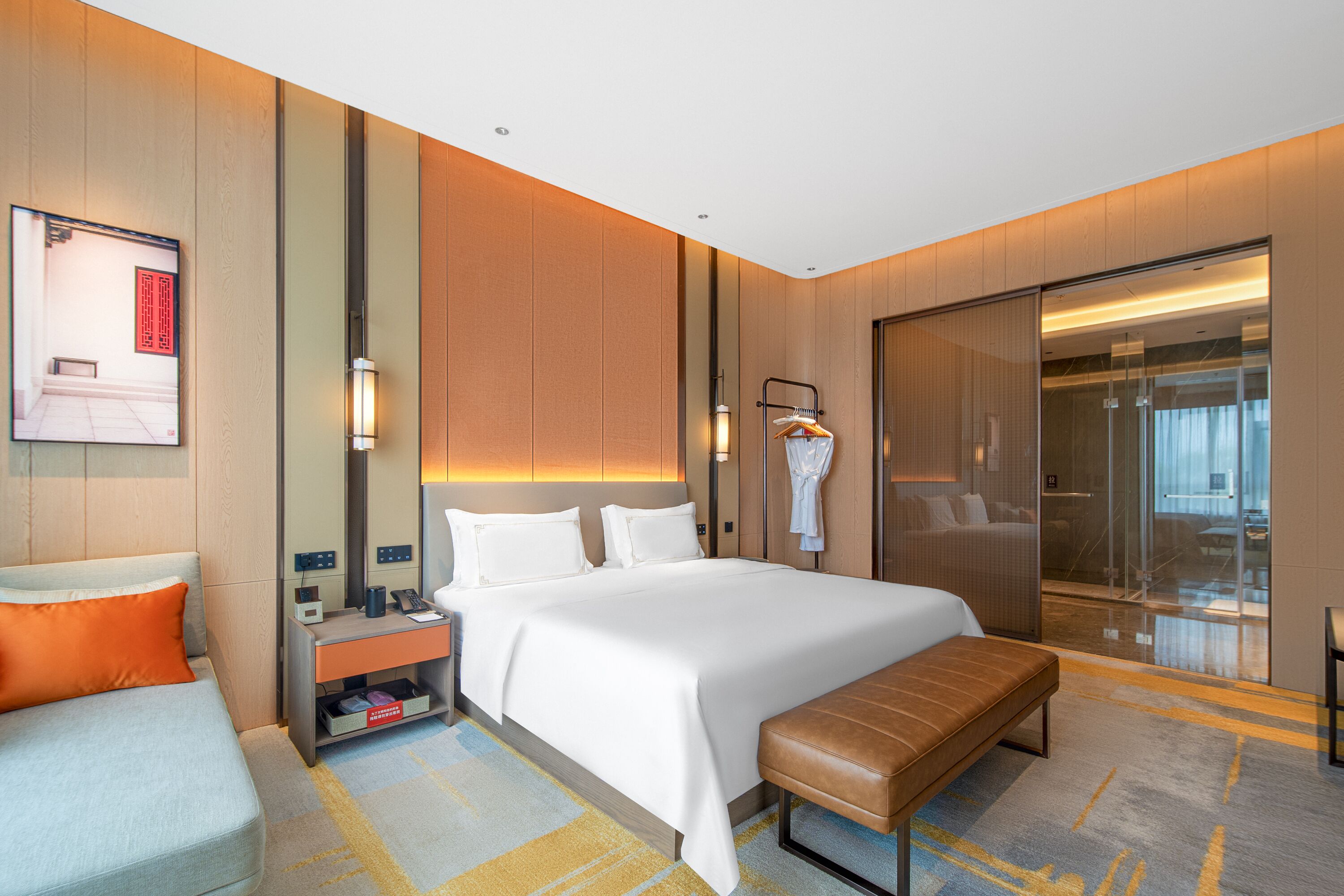 A hotel room with a large bed, a bedside table, a bench, a wardrobe, and a sliding door leading to a bathroom.