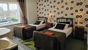 A hotel room with two beds, a window with curtains, a radiator, a small table, and a green rug.