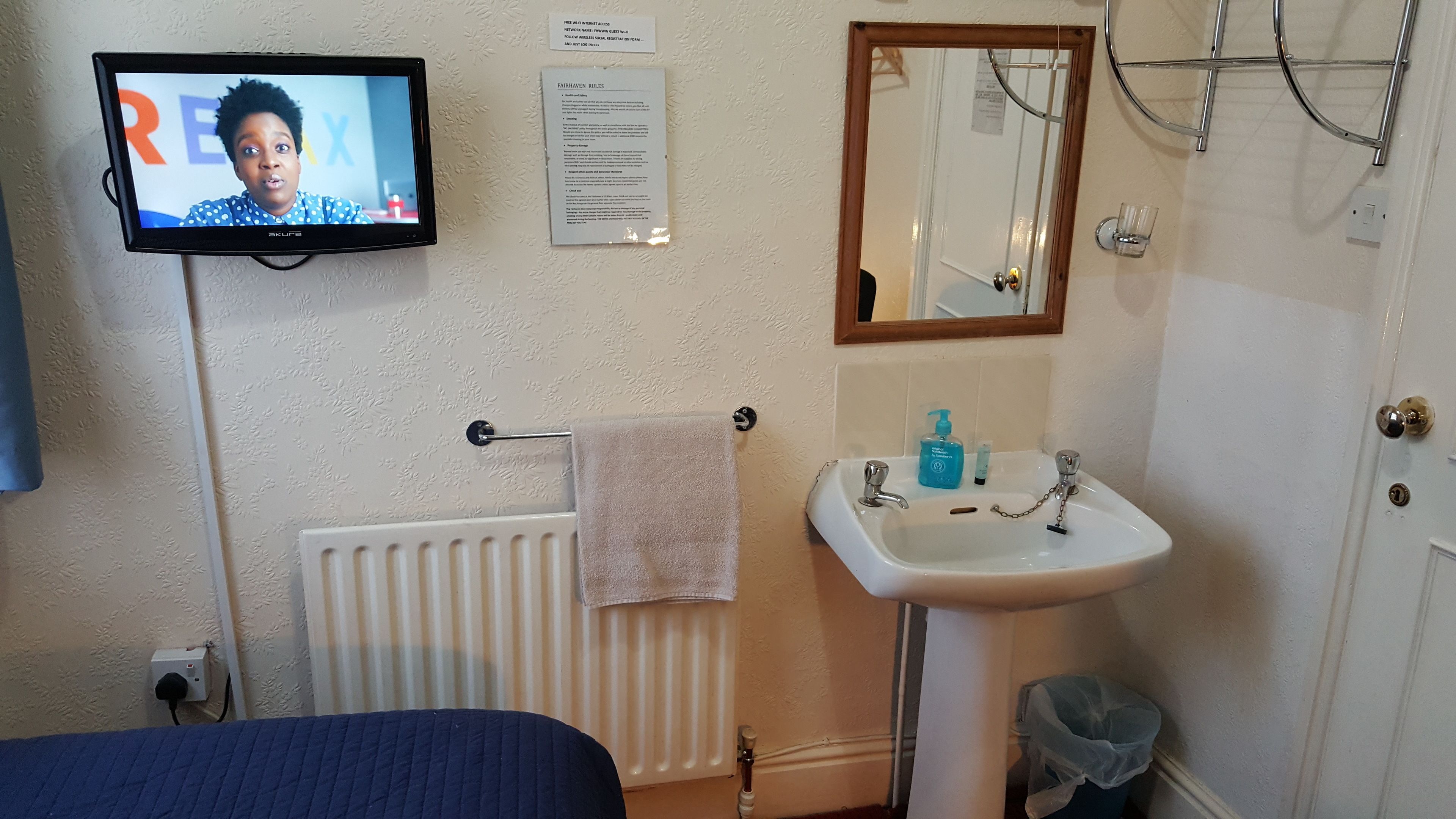 Ein Badezimmer mit Waschbecken, Spiegel, Handtuchhalter und einem Fernseher, der eine Person zeigt.