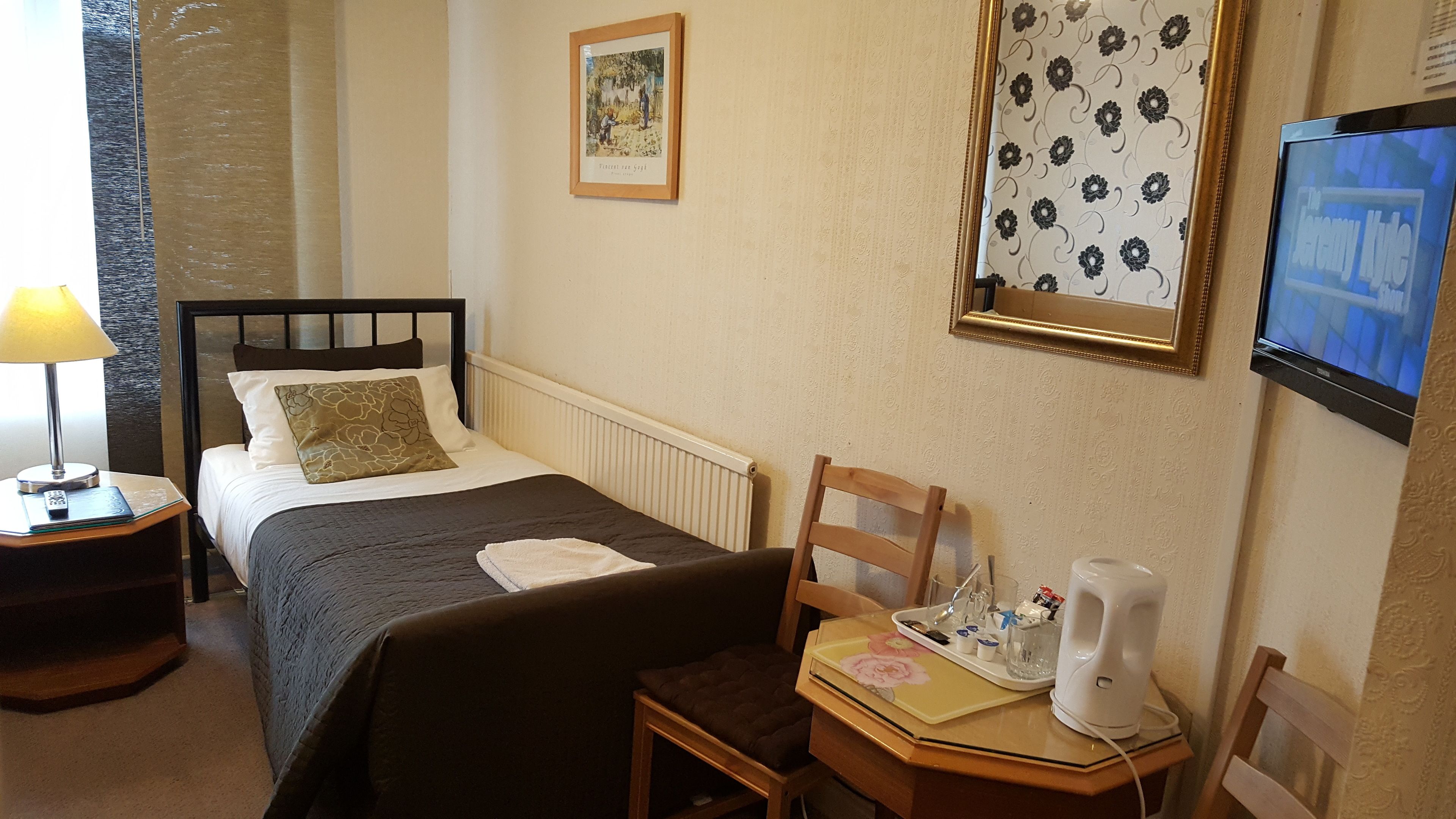 A hotel room with a bed, a bedside table with a lamp, a chair, a small table with a tray of toiletries, a framed picture, and a TV mounted on the wall.