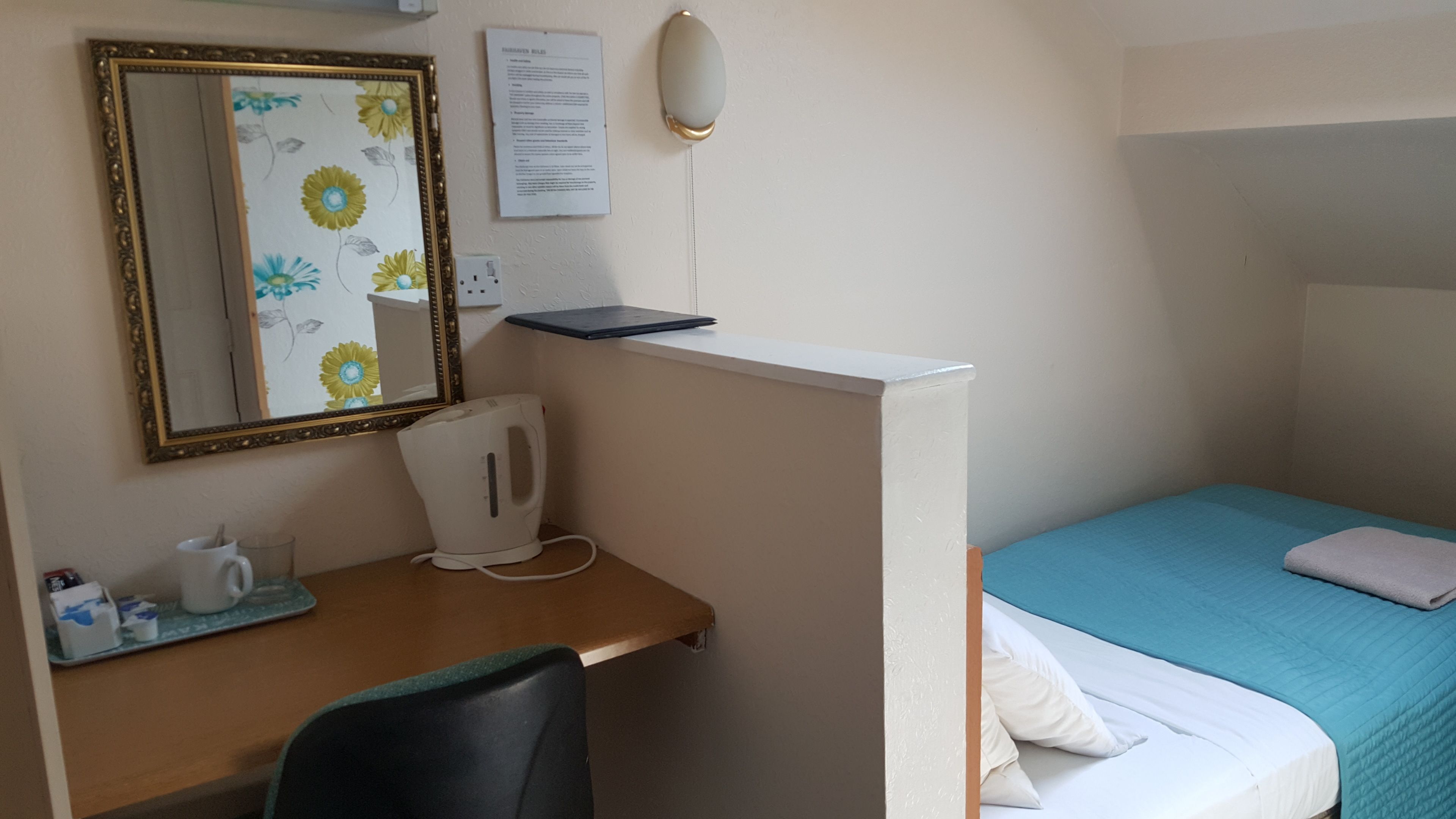 A small, neatly arranged room with a desk, chair, and a bed with a blue bedspread.