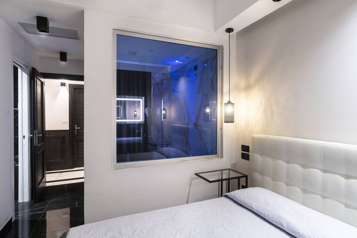 Une chambre d’hôtel moderne avec une grande fenêtre, un lit doté d’une tête de lit blanche, une table en verre et un luminaire fixé au mur.