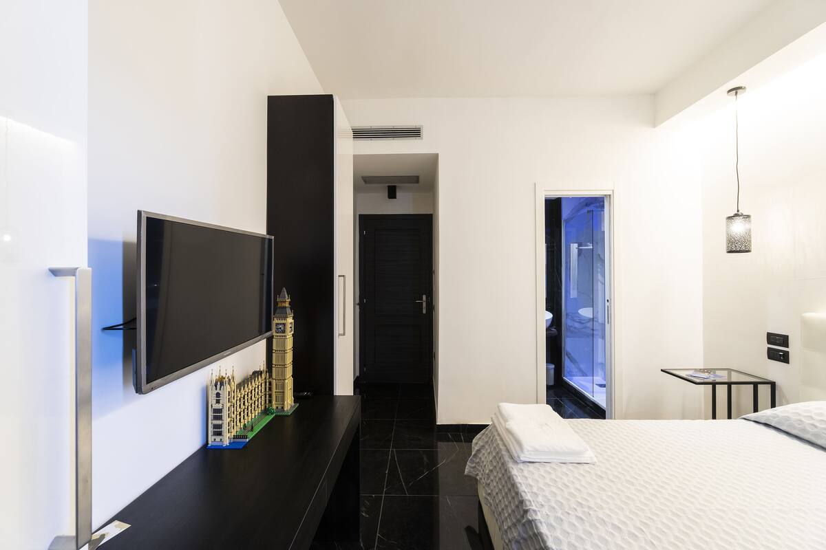 Une chambre d’hôtel moderne équipée d’un téléviseur à écran plat fixé au mur, d’un lit recouvert d’une couette blanche, d’une petite table avec une lampe et offrant une vue sur la ville par la fenêtre.