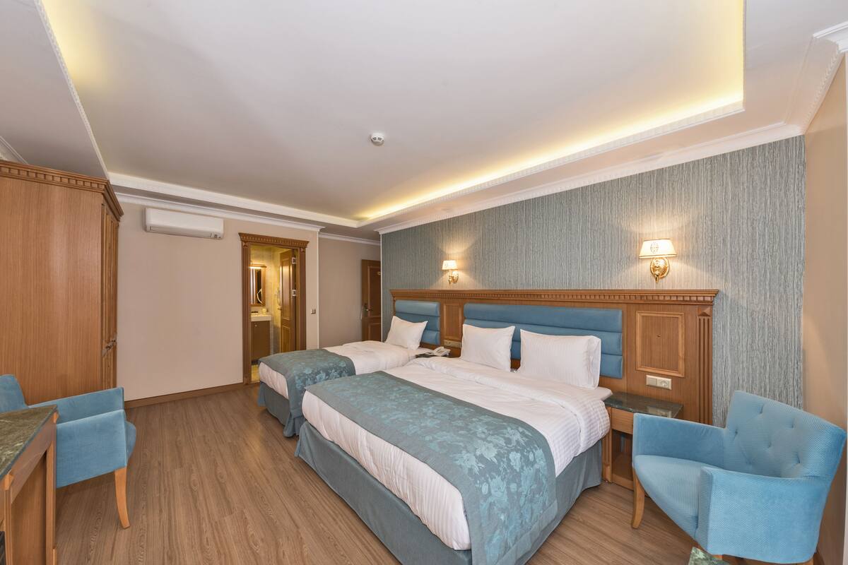 Habitación de hotel con dos camas, un escritorio, una silla y un baño visible a través de una puerta abierta.
