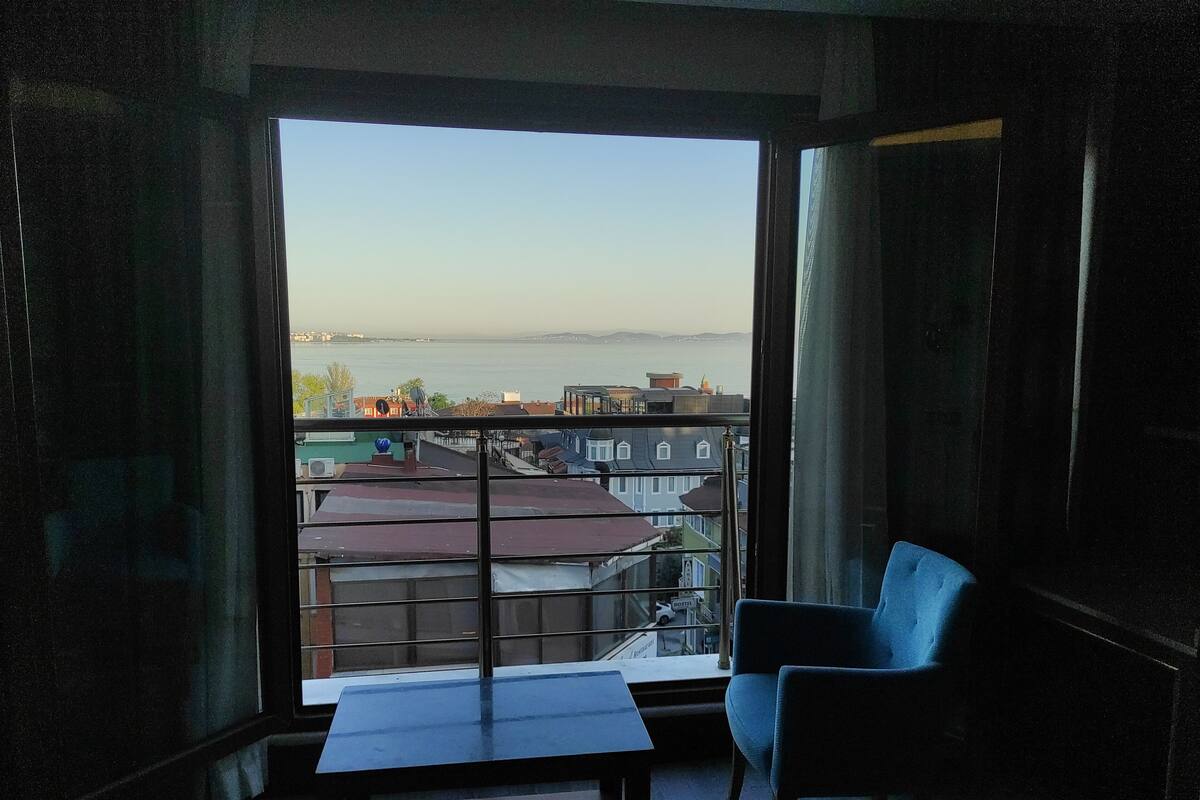 Habitación con una ventana grande que da a vistas de la ciudad y al agua. Hay una mesa y una silla dentro.