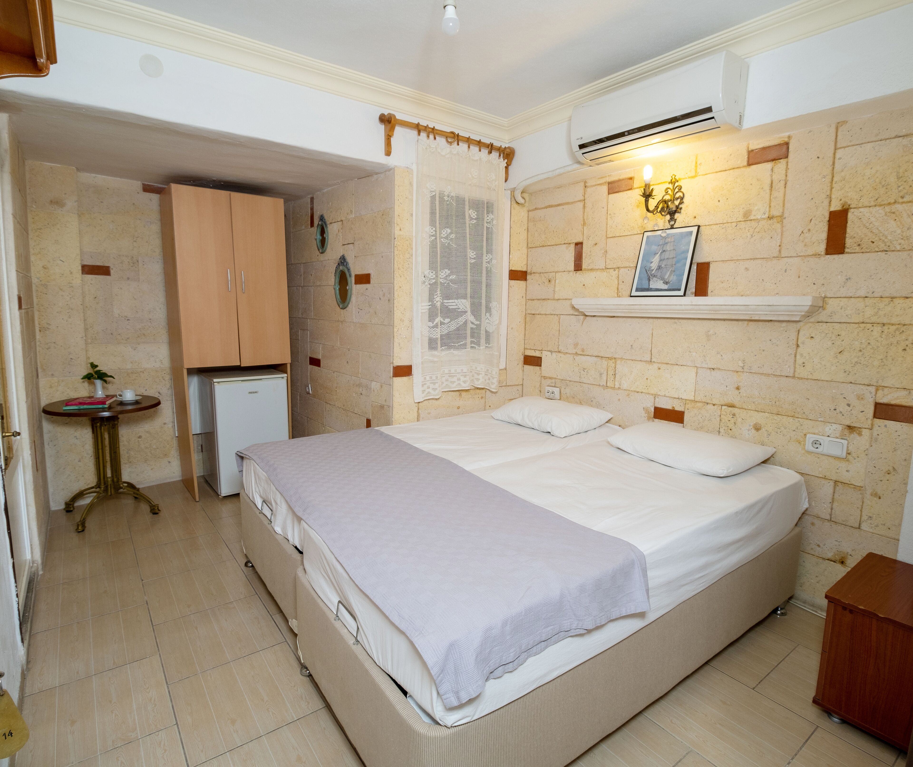 Une chambre avec un lit, une petite table sur laquelle est posée une plante, une étagère fixée au mur avec un tableau, et un appareil de climatisation.