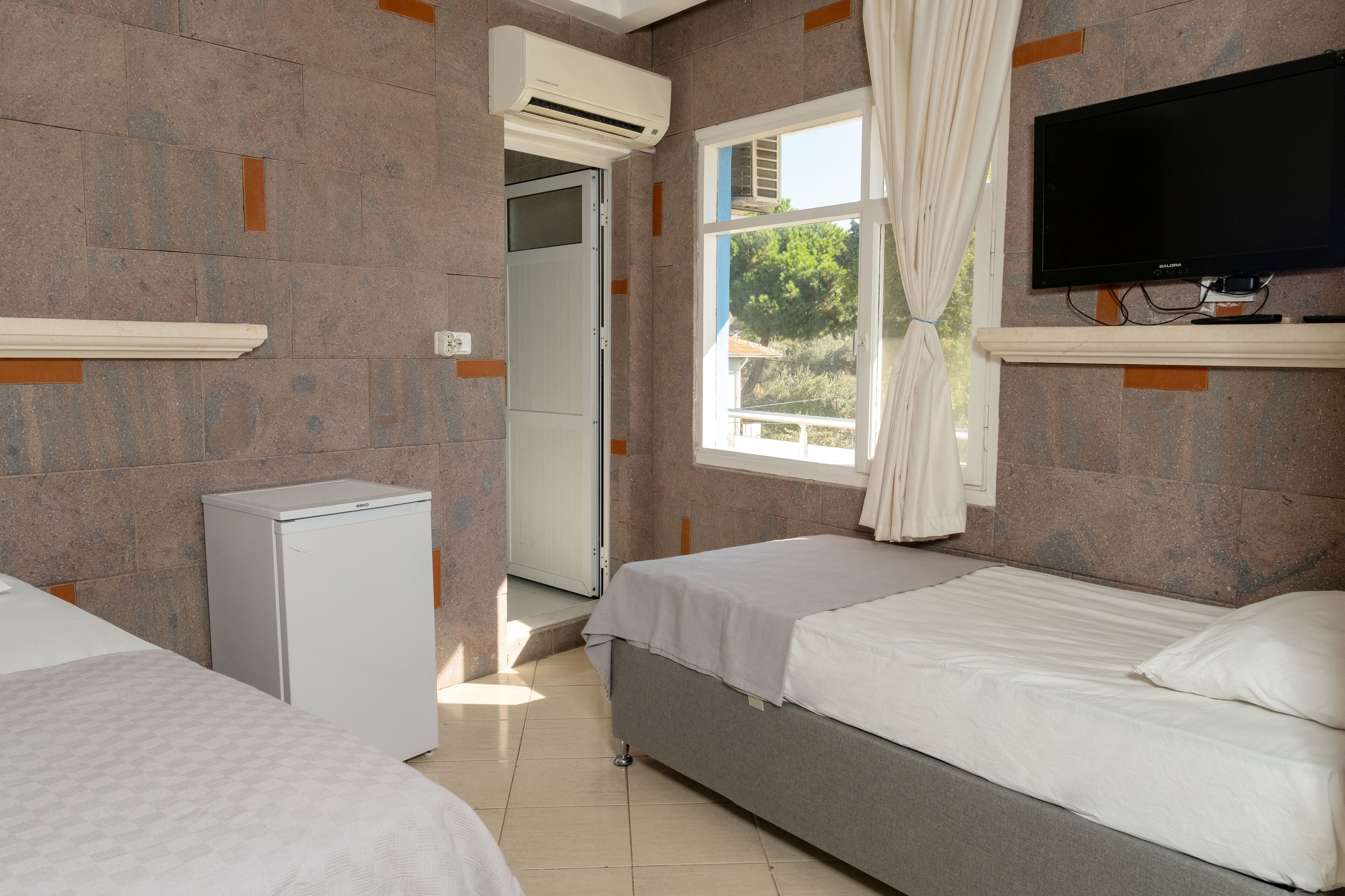 A hotel room with two beds, a TV, a mini-fridge, and a window with curtains.