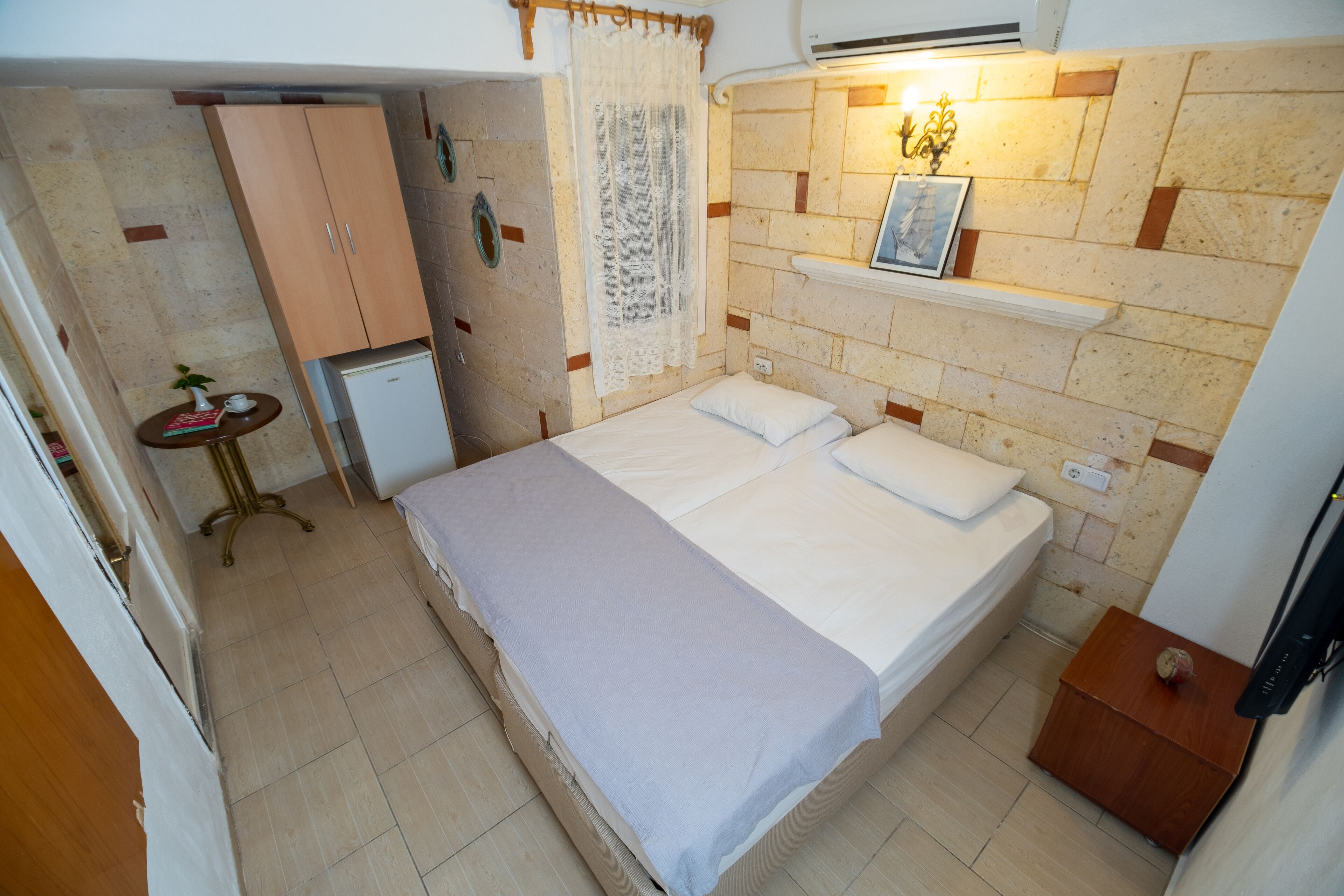A small, neatly arranged bedroom with a bed, a small table with a plant, a wardrobe, and a wall-mounted shelf.