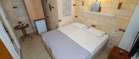 Une petite chambre à coucher, bien rangée, avec un lit, une petite table sur laquelle est posée une plante, une armoire et une étagère fixée au mur.