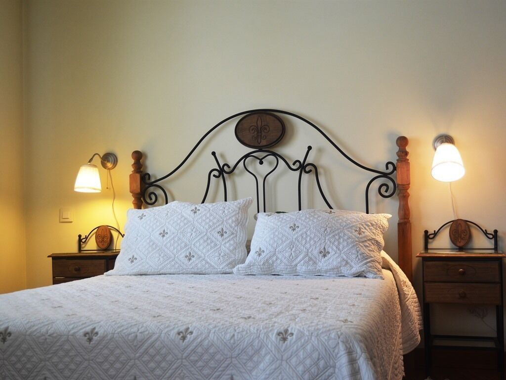 Cama bem arrumada com edredom branco acolchoado, duas almofadas combinando e cabeceira de metal com uma decoração em relevo. Há duas mesas de cabeceira com abajures de cada lado da cama.
