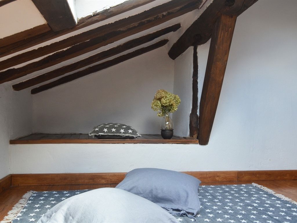 A small, cozy room with a sloped ceiling, wooden beams, a window, a shelf with a potted plant, and a bed with star-patterned bedding.
