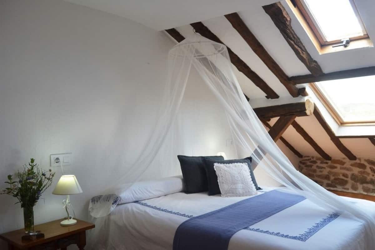 Een slaapkamer met een bed onder een wit baldakijn, een nachtkastje met een lamp en een uitzicht op de lucht door een dakraam.
