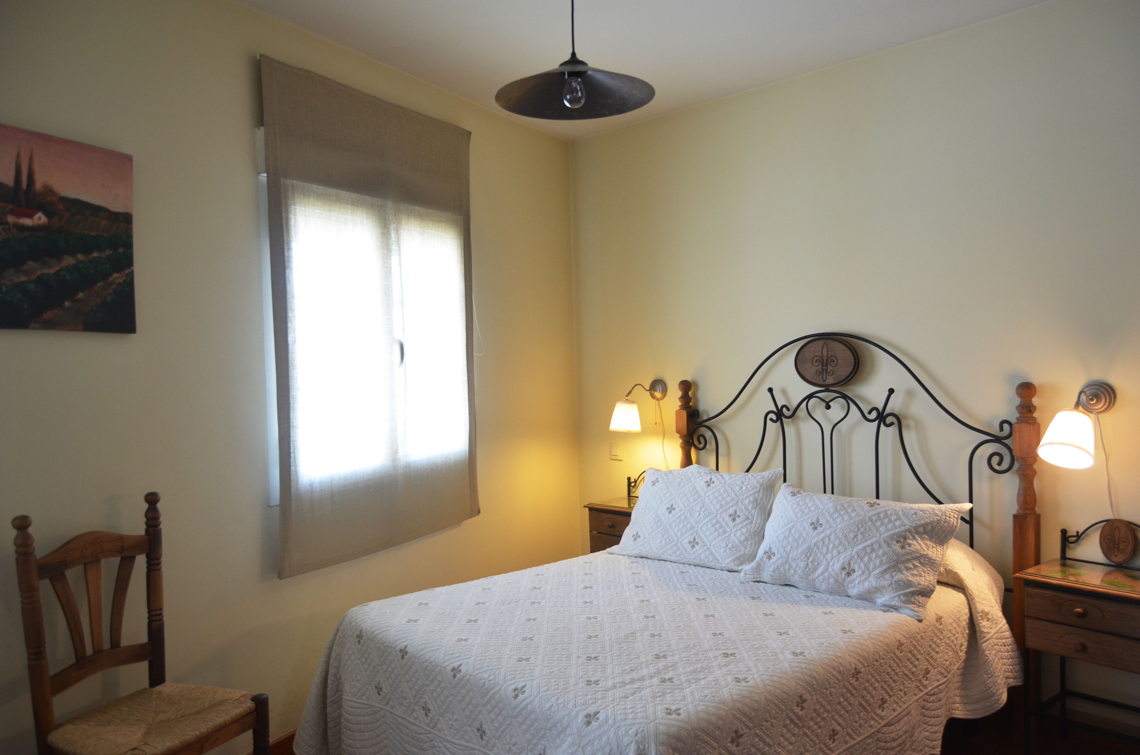 Un dormitorio con cama, dos lámparas de noche, una silla de madera, una mesita de noche y una ventana con persianas.