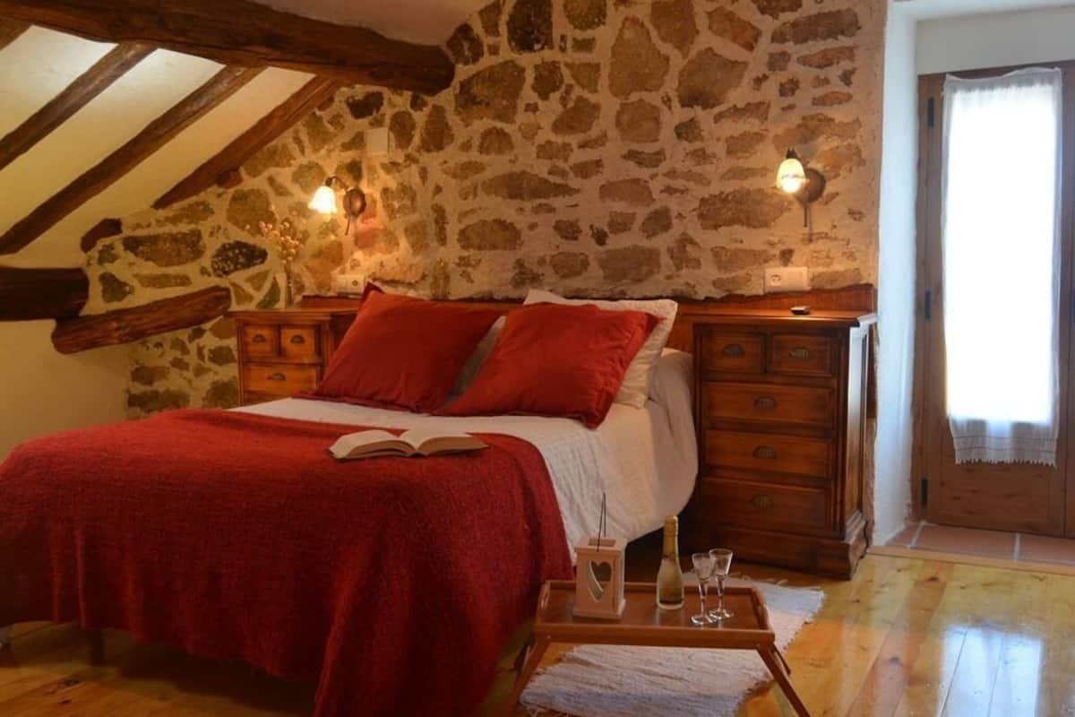 Een slaapkamer met een stenen muur, houten balken, een bed met rode beddengoed, een houten dressoir en een nachtkastje met een fles en glazen.