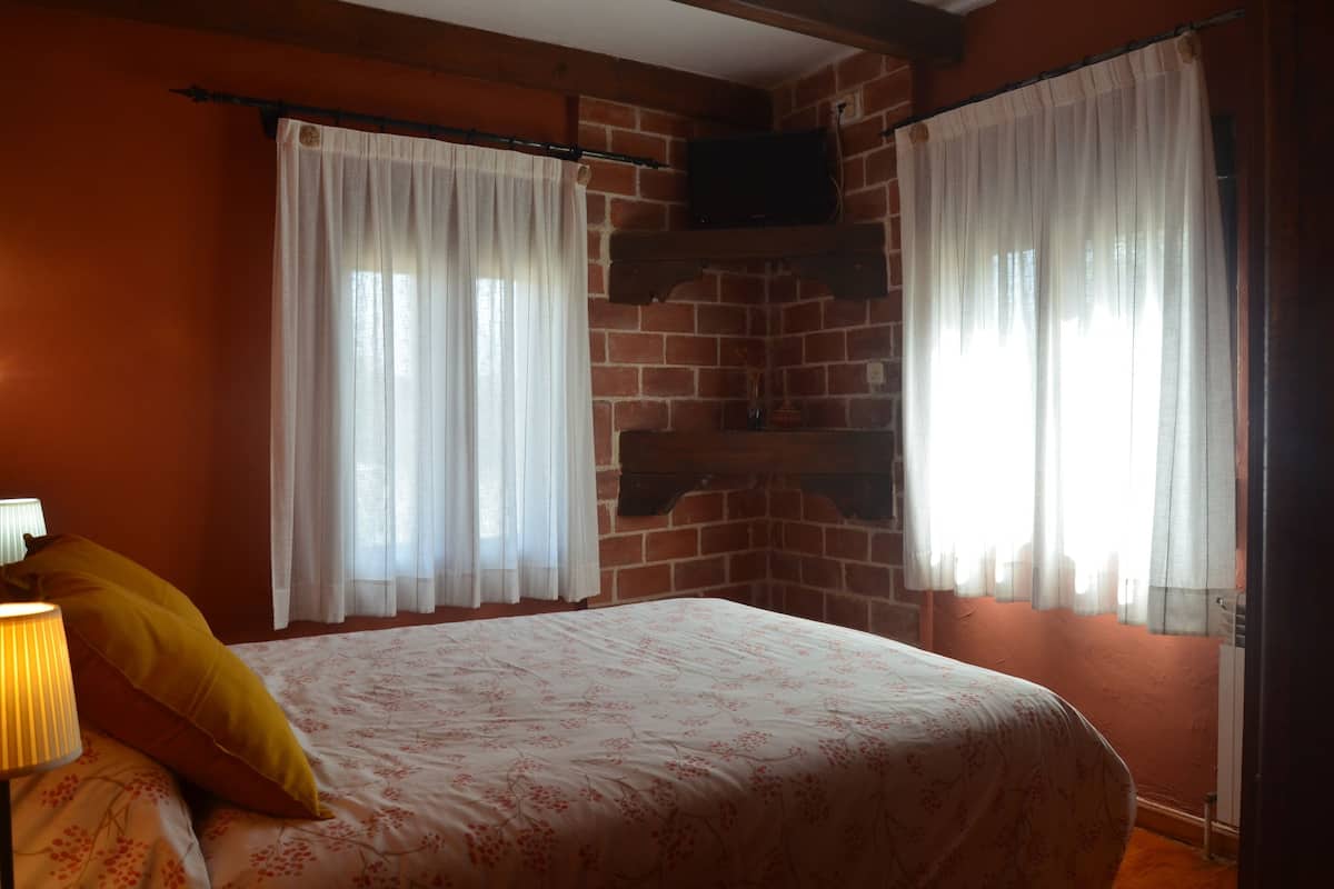 Een slaapkamer met een bed, twee ramen met gordijnen, een wandgemonteerde televisie en een bakstenen muur.
