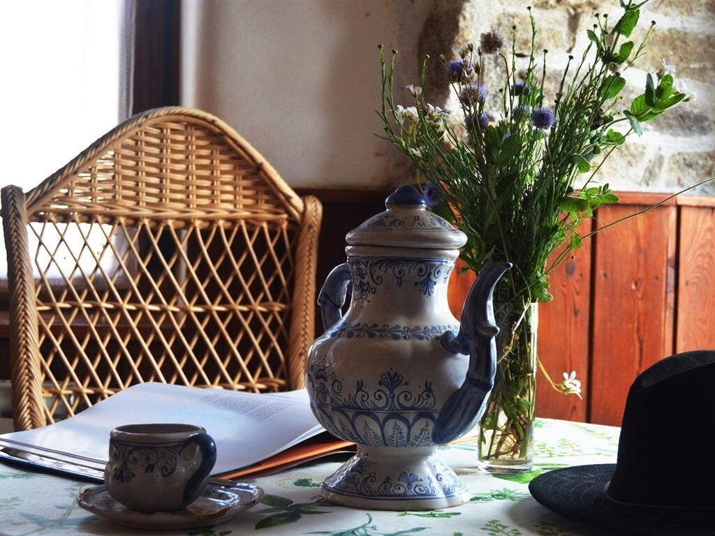 Una silla de mimbre, una mesa con mantel floral, una jarra de cerámica, un florero con flores y un sombrero negro.