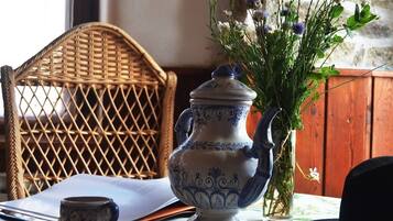 Een rieten stoel, een tafel met een bloemrijk tafelkleed, een keramieken kan, een vaas met bloemen en een zwarte hoed.