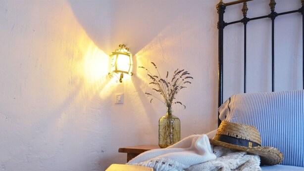 A cozy room with a bed, a bedside table with a lamp, a vase with dried plants, a straw hat, a book, and a blanket. - El Refugio de Gredos - Hostel (Navarredonda de Gredos)