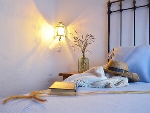 Una habitación acogedora con una cama, una mesita de noche con una lámpara, un jarrón con plantas secas, un sombrero de paja, un libro y una manta.