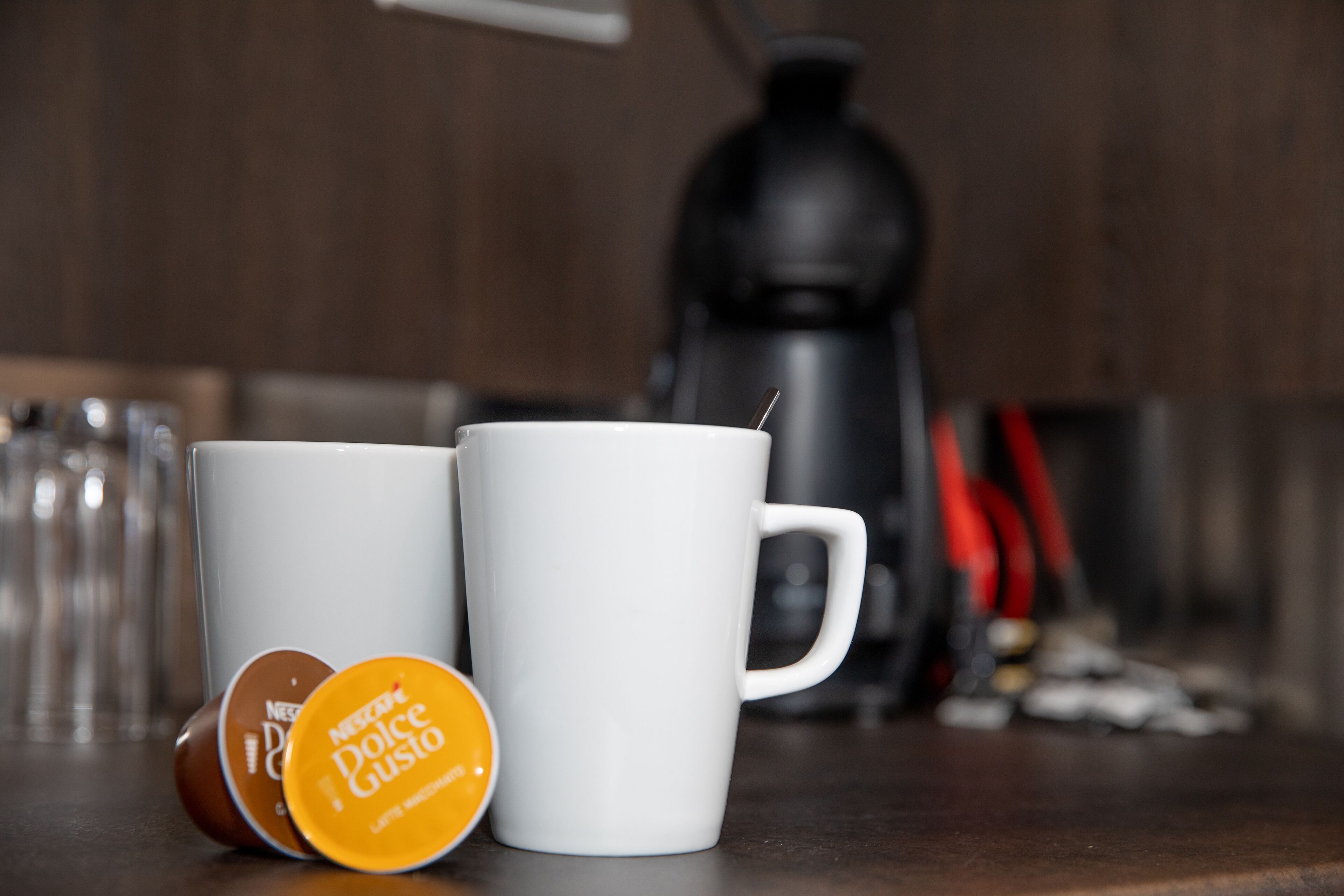 Two white mugs on a kitchen counter with a coffee maker in the background.