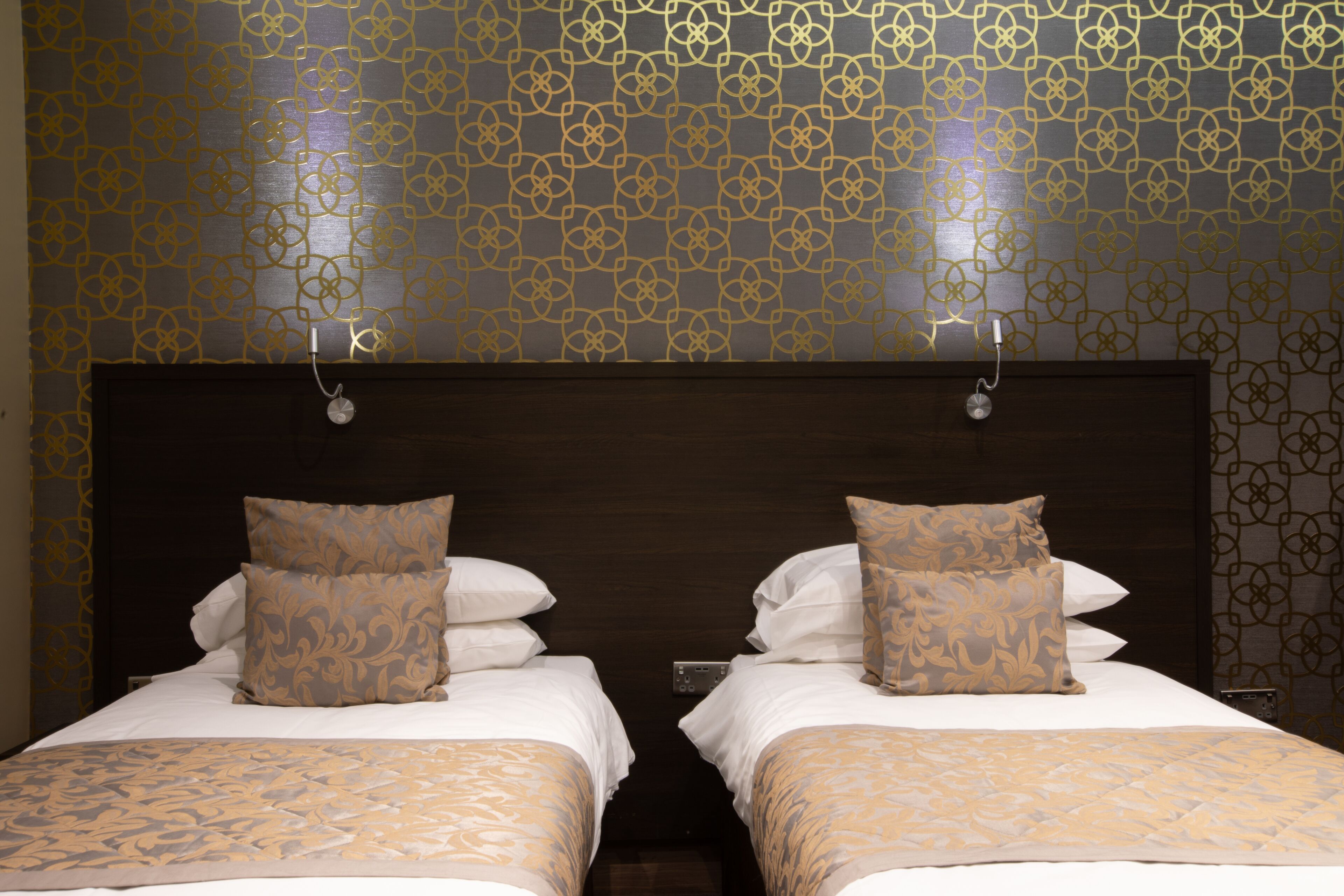 Ein Hotelzimmer mit zwei Betten, jeweils mit gemusterten Bettbezügen und Kissen, einem dunklen Holz-Kopfende und einer Wand mit geometrischem Muster.