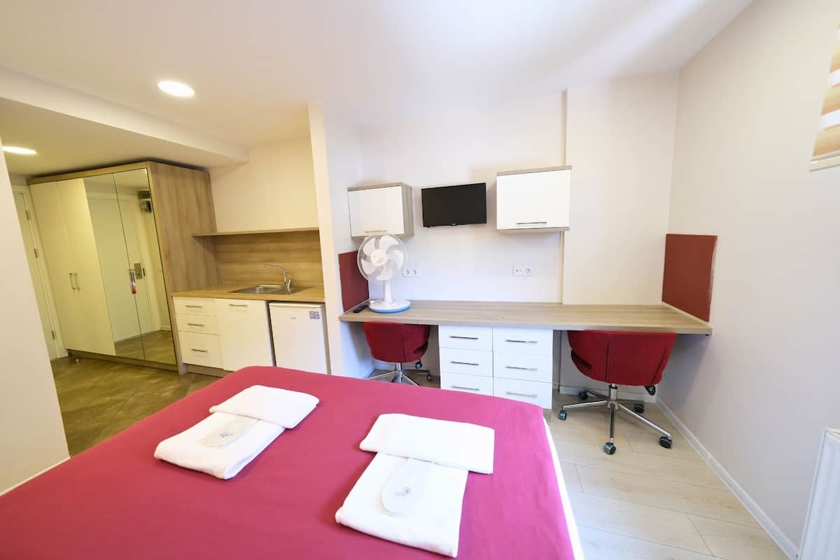 A hotel room with a bed, a desk, a chair, a TV, a fan, and a kitchenette.