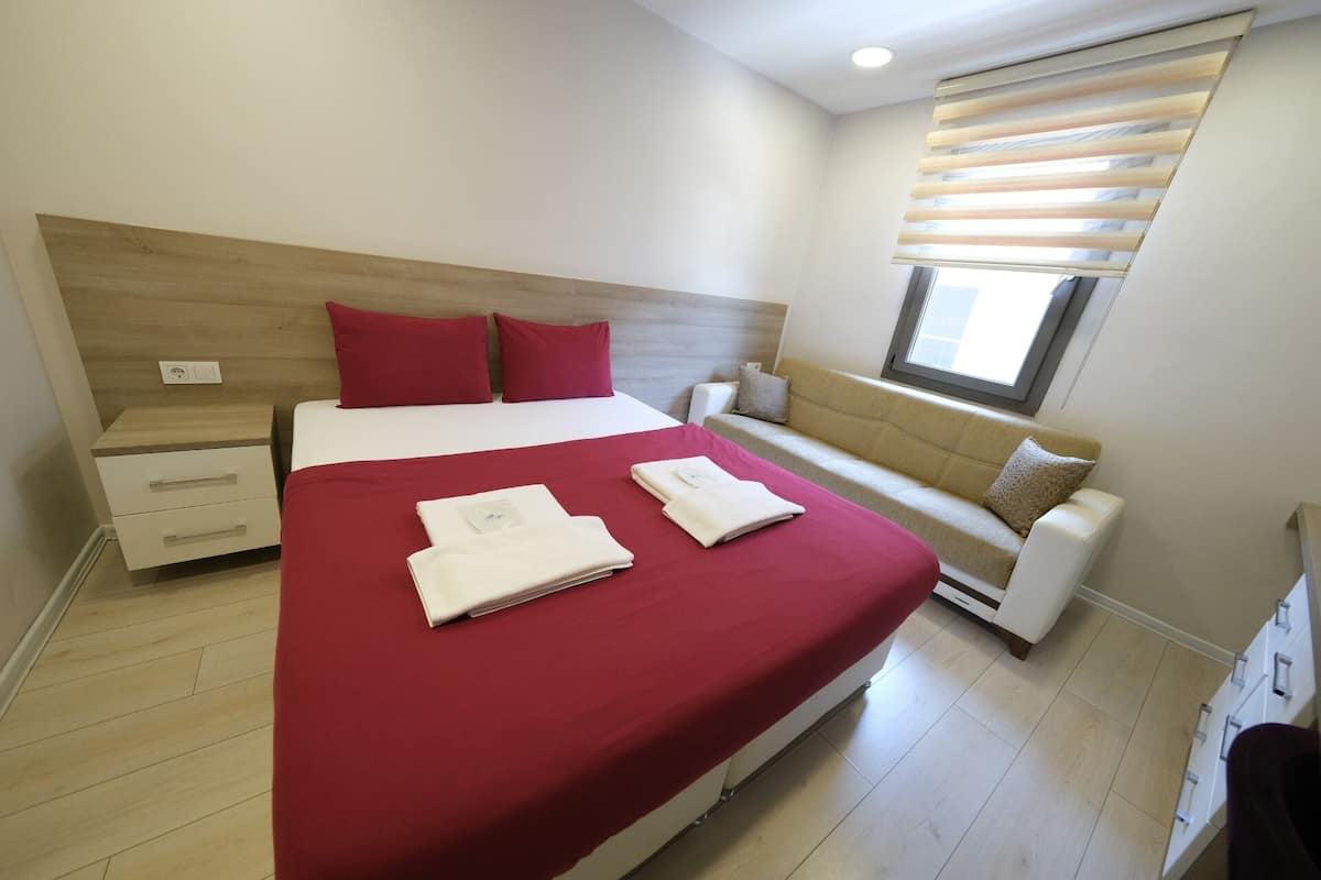 A bedroom with a bed, a bedside table, a sofa, and a window with blinds.