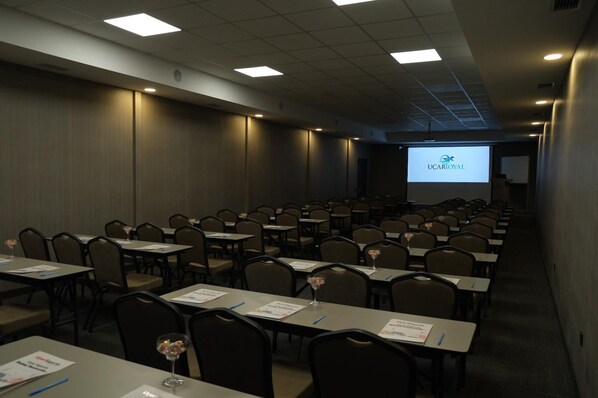 Un salón de conferencias con filas de mesas y sillas, una pantalla de proyector mostrando 'UCAR ROYAL' y un atril para presentaciones.
