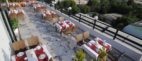 An outdoor terrace with tables set for dining, surrounded by greenery and overlooking a cityscape.