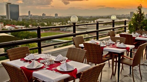 Restaurante com mesas preparadas para refeições, com vista para a paisagem urbana ao pôr do sol.