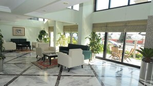 Um hall amplo com piso de mármore, área de estar com sofás e cadeiras, grandes janelas e plantas em vasos.