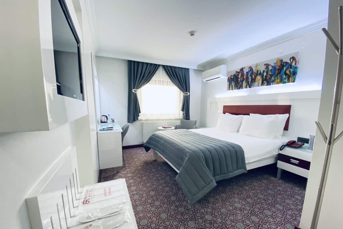 A hotel room with a bed, a desk, a chair, a window with curtains, a painting on the wall, and a patterned carpet.
