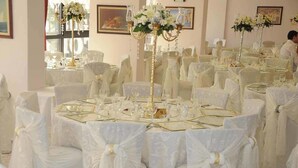 Uma sala de banquetes com mesas redondas, toalhas brancas e cadeiras adornadas com fitas brancas.