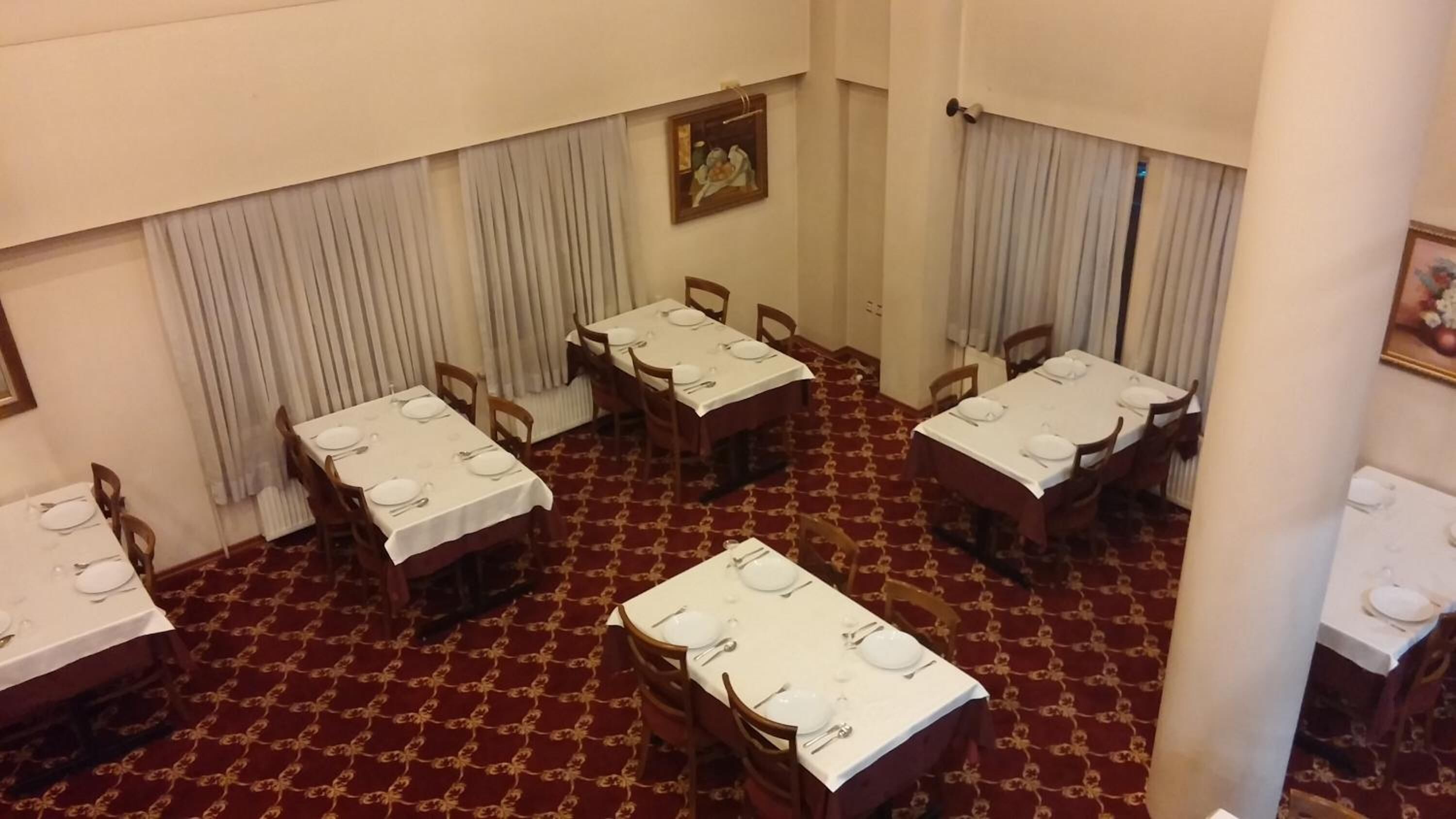 Un comedor con cuatro mesas, cada una con mantel blanco y cubiertos. La habitación tiene una alfombra estampada y cortinas blancas.