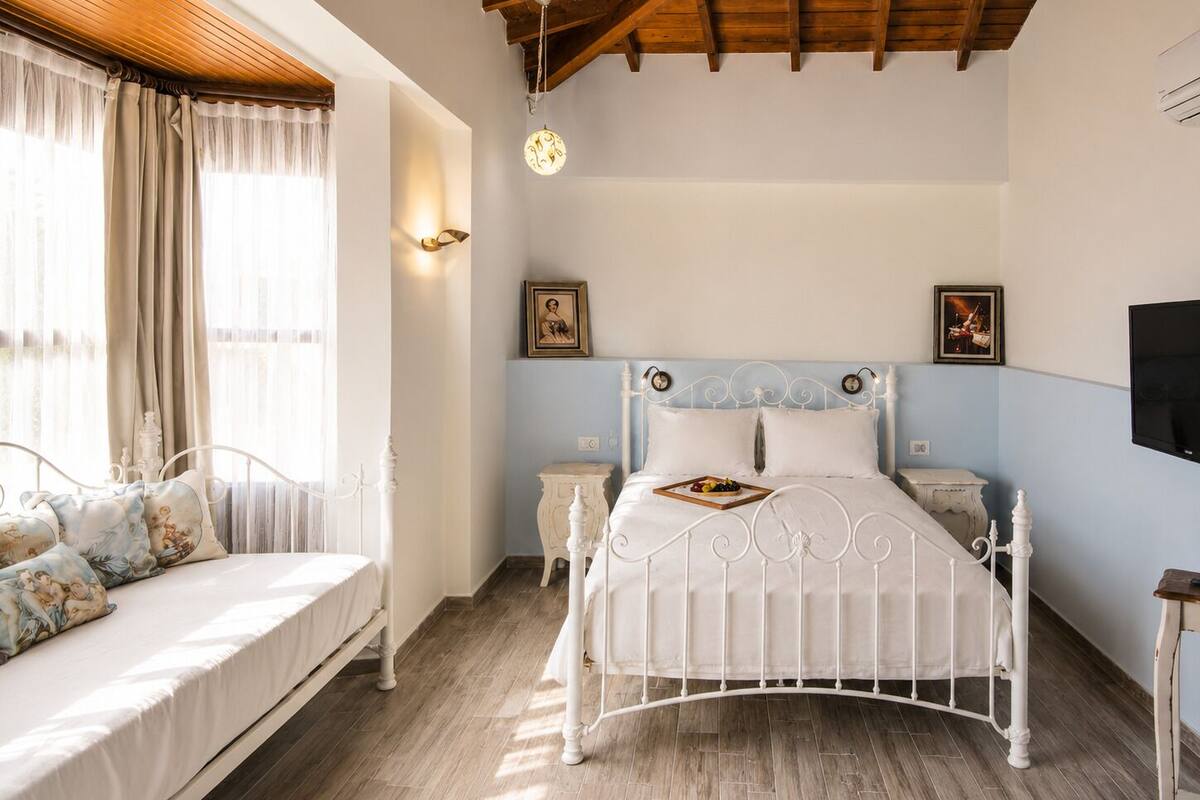 A bedroom with a white metal bed, a bedside table, a nightstand, a wall-mounted TV, and a window with curtains.