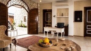 Quarto com parede de pedra, porta de madeira, escrivaninha, computador e mesa redonda de madeira com frutas.