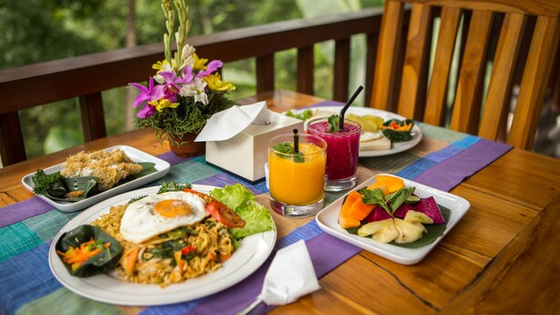A wooden table with a variety of dishes, including fried rice, noodles, and vegetables, along with two glasses of juice. - Ayu Duwur Beji (Ubud)