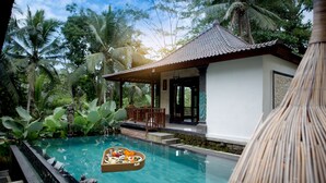 A serene pool area with a floating basket of food, surrounded by lush greenery and a traditional building with a tiled roof. - Ayu Duwur Beji (Ubud)