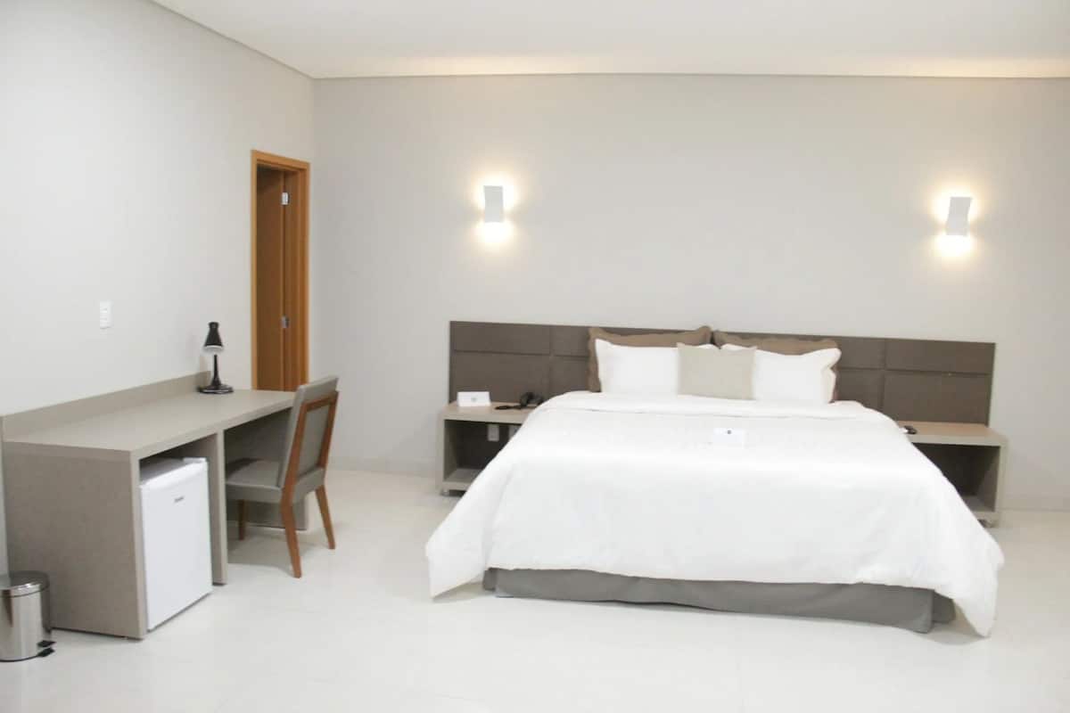 Quarto de hotel com uma cama, uma escrivaninha, uma cadeira e um pequeno frigorífico.