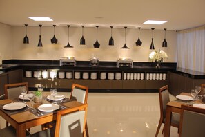 Un comedor moderno de hotel con una larga barra, varias mesas y lámparas colgantes.