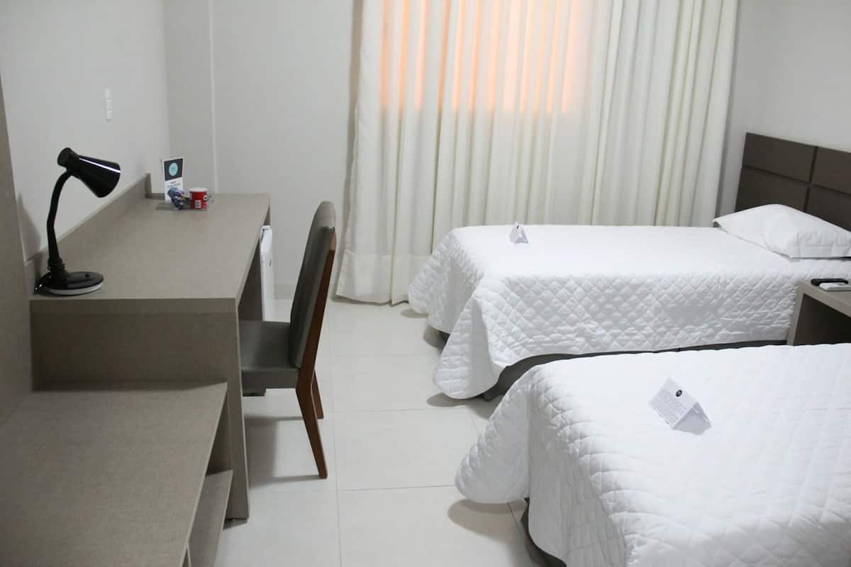 Quarto de hotel com duas camas, uma escrivaninha com abajur, uma cadeira e uma janela com cortinas.